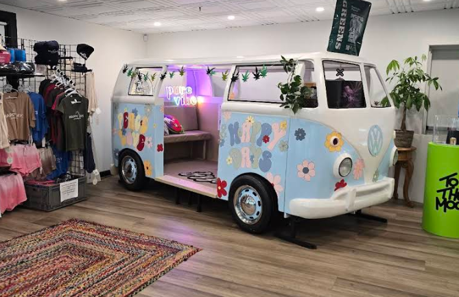 Interior of a retail boutique featuring a retro Volkswagen van painted white and light blue with flower decals, decorated with cannabis leaf garlands and a 'pure vibe' neon sign inside. The left side displays branded apparel, including hats and colorful shirts, while the right side features a green cylindrical pedestal with 'To the Moon' branding and a potted Money Tree. The space has a vintage-inspired 'Peace & Love' theme with bright lighting and a braided multicolor rug on the wooden floor