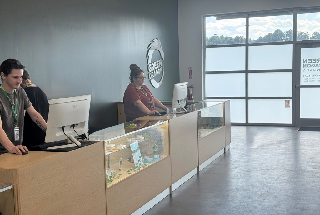Interior of Green Dragon Yulee cannabis dispensary with staff behind wooden counters, glass product display cases, computer monitors, and Green Dragon branding on a dark accent wall near large front windows