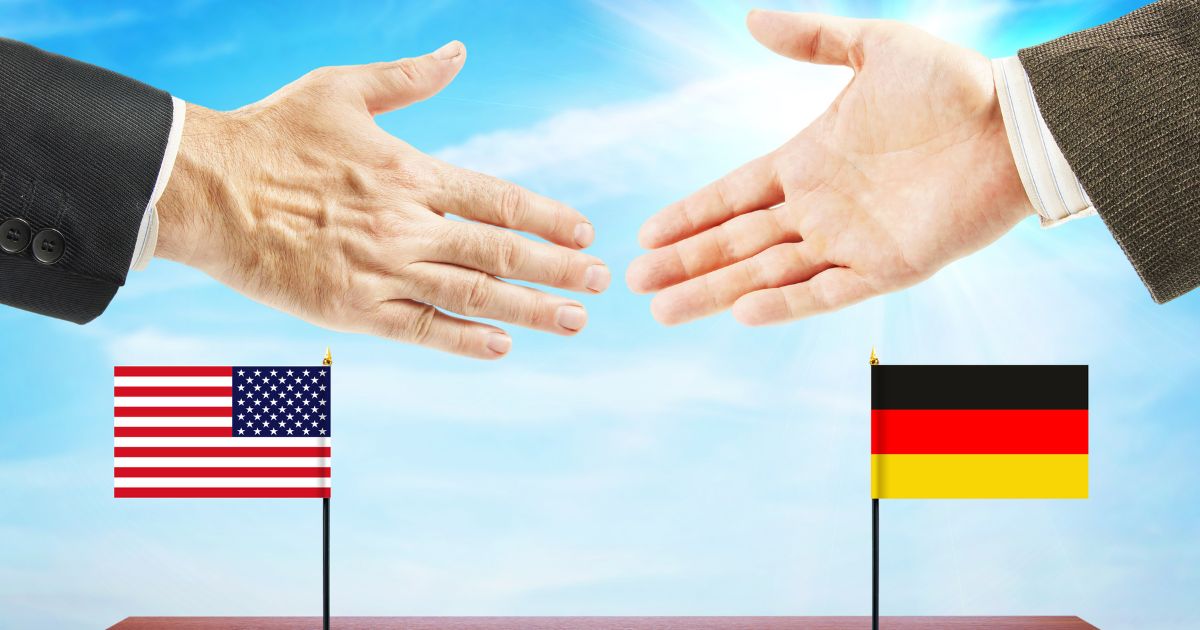 Two business-clad hands reach toward a handshake above small U.S. and German flags against a bright blue sky, symbolizing Germany cannabis reform and international policy comparison