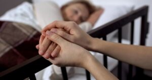 Close-up of two hands gently holding an older patient’s hand beside a bed, with a woman resting in soft focus in the background, illustrating compassionate end-of-life care under Ryan’s Law.