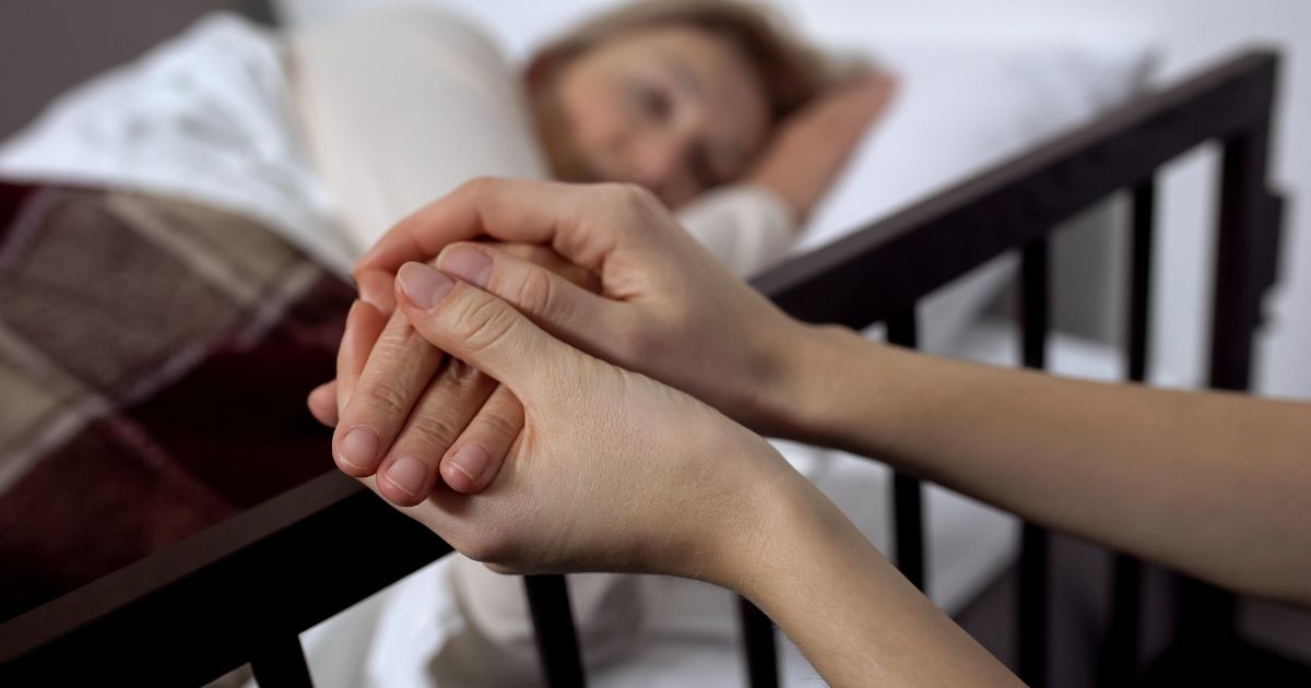 Close-up of two hands gently holding an older patient’s hand beside a bed, with a woman resting in soft focus in the background, illustrating compassionate end-of-life care under Ryan’s Law.