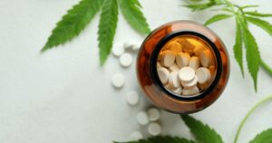 Medical cannabis leaves, an open amber bottle, and scattered white pills rest on a white textured surface.