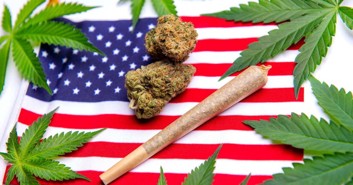 Top-down view of cannabis buds, a rolled joint, and fresh green leaves arranged on an American flag, symbolizing American cannabis legalization.