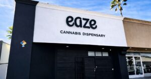 Exterior of the Eaze cannabis dispensary in Van Nuys, California, featuring a minimalist design with bold black 'eaze' branding and 'CANNABIS DISPENSARY' text on a white facade. The storefront includes a black roll-up door, a safety NFPA 704 diamond sign, and palm trees against a clear blue sky