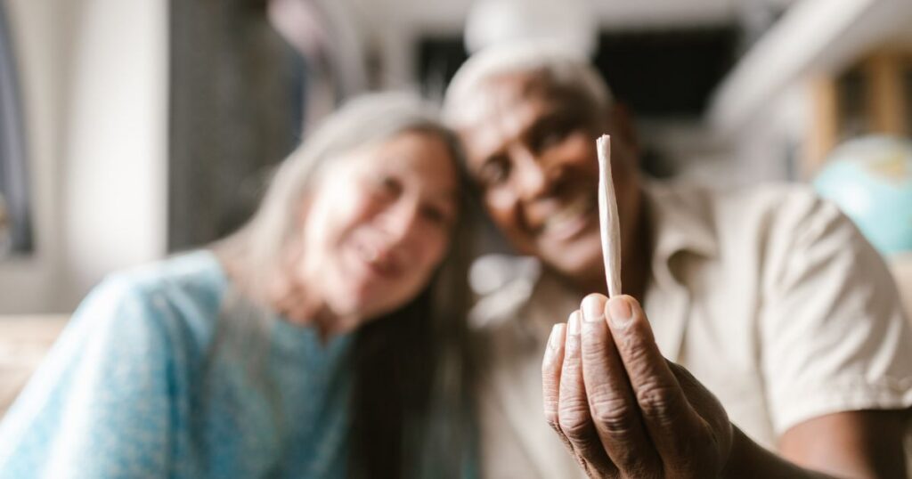 Close-up of a hand holding a hand-rolled joint with a smiling couple blurred indoors, highlighting older adults cannabis use.