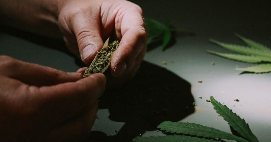 "Close-up of hands rolling a small cannabis joint on a dark surface, surrounded by scattered cannabis leaves and loose plant material, illustrating the cannabis micro dose trend