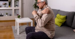 Older man sitting on a sofa and clutching his shoulder in pain in a bright living room, illustrating a men’s health concern related to chronic conditions and wellness.
