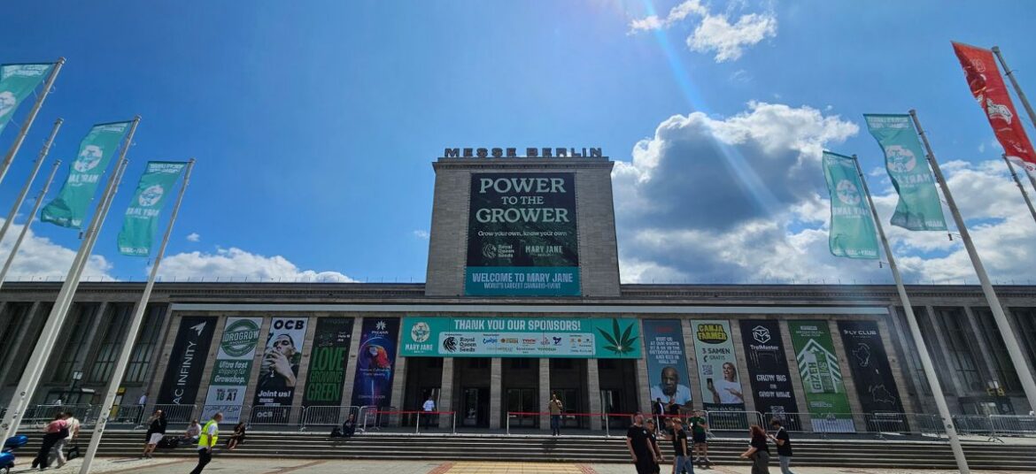 Entrance to Messe Berlin decorated with large event banners, sponsor signage, and flags for Mary Jane Berlin 2026, with visitors walking across the plaza under a bright blue sky.
